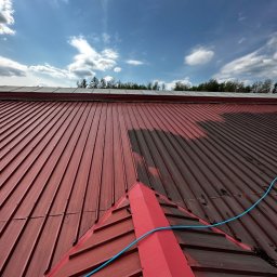 Rydel - Mycie Wysokociśnieniowe - Częściowo umyty, czerwony dach z blachy trapezowej, widoczny niebieski wąż do mycia pod ciśnieniem, błękitne niebo z chmurami w tle.