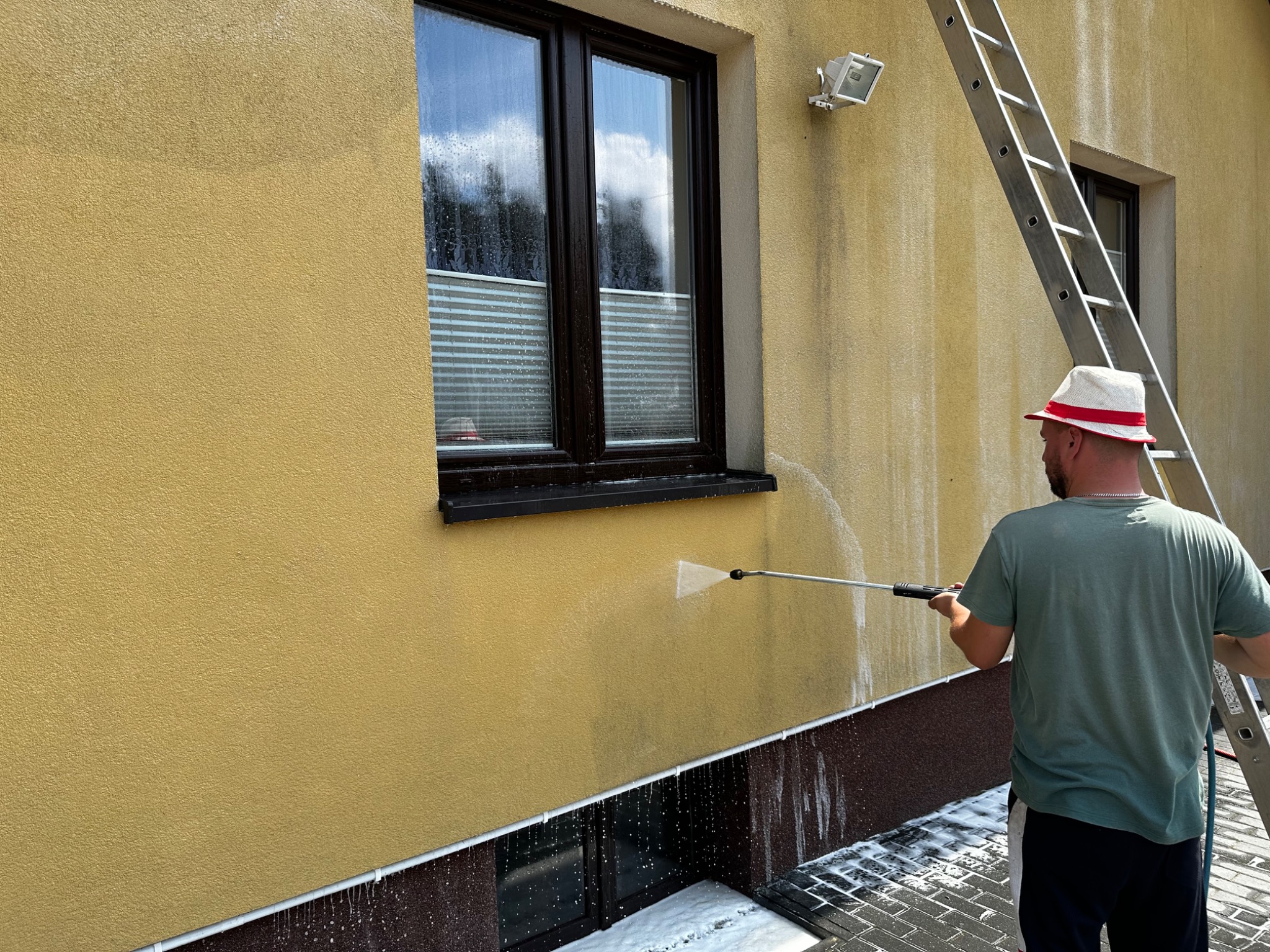Czyszczenie elewacji budynku z żółtym tynkiem za pomocą myjki ciśnieniowej; widoczna drabina oparta o ścianę i mężczyzna w kapeluszu.