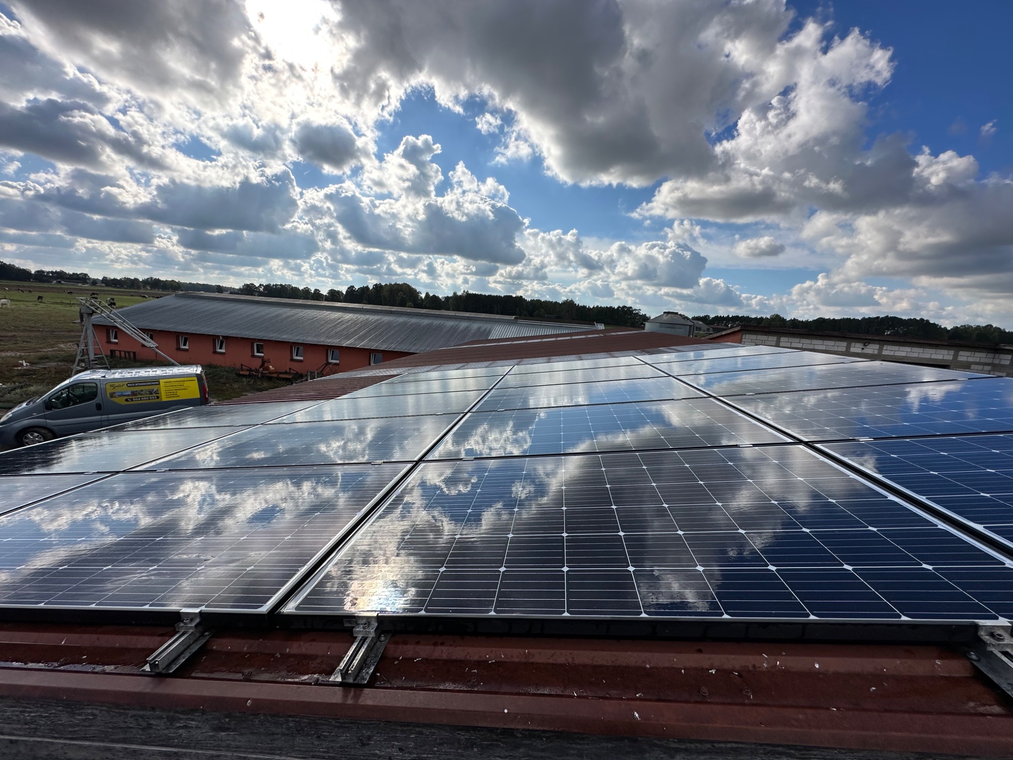 Błękitne niebo z chmurami odbija się w panelach słonecznych zamontowanych na czerwonym dachu budynku gospodarczego, z widocznym samochodem dostawczym w tle.