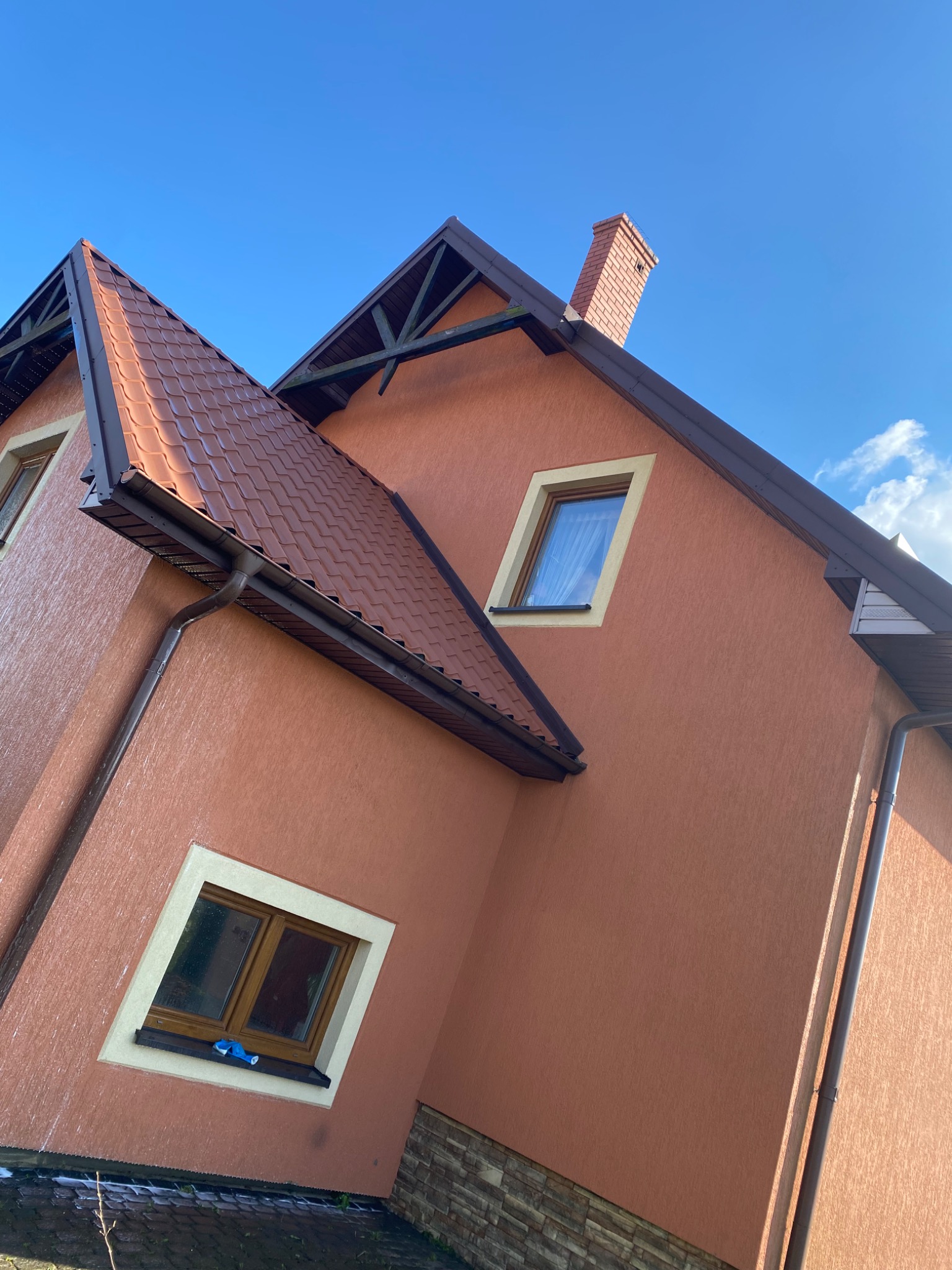Fragment elewacji domu z brązowym dachem i rynnami, widoczny komin i okna z jasnymi obramowaniami na tle błękitnego nieba.