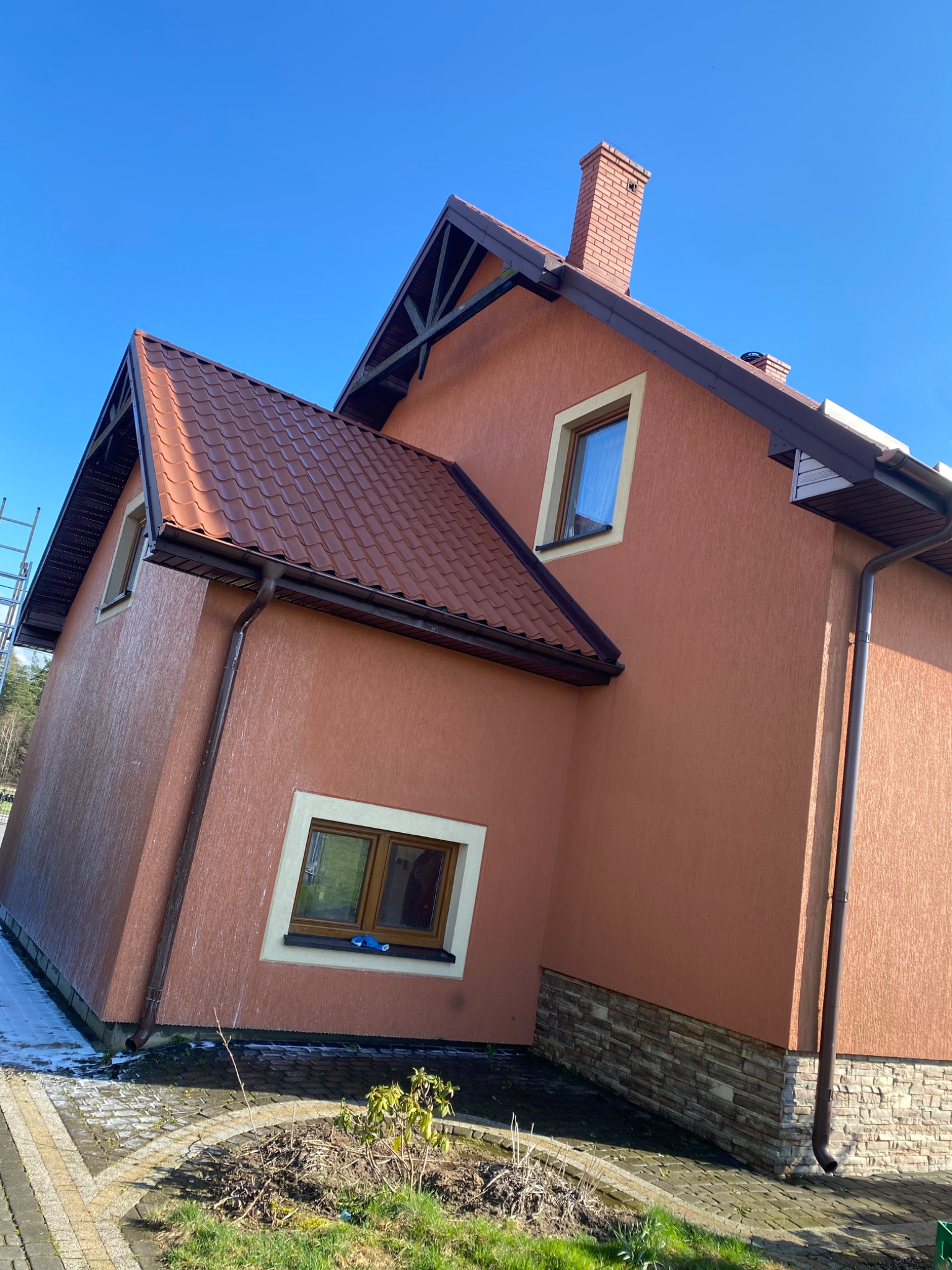 Dwupiętrowy dom z pomarańczową elewacją, brązowym dachem z dachówki i ceglanym kominem, widziany z boku w słoneczny dzień, z widoczną drabiną opartą o ścianę.