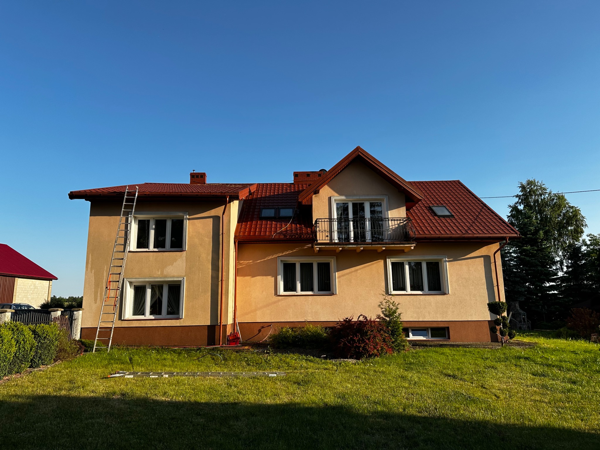 Dwupiętrowy dom z beżową elewacją i czerwoną dachówką, z przystawioną drabiną aluminiową, sugerującą prace na wysokości.