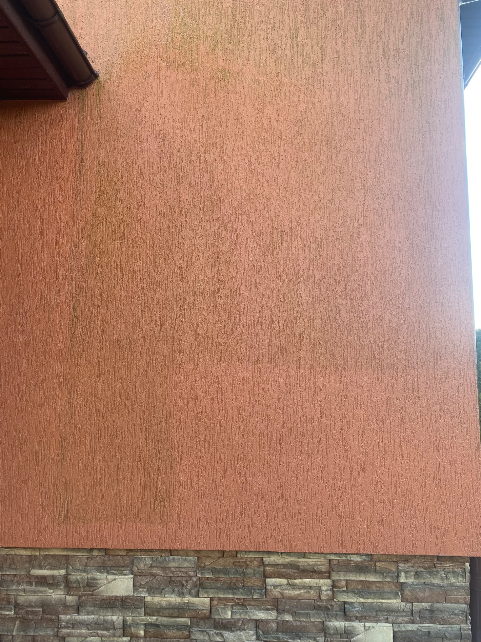 Fragment ściany budynku z elewacją w kolorze terakoty z widocznymi śladami zabrudzeń i zielonego nalotu, poniżej wykończenie z płytek imitujących kamień.