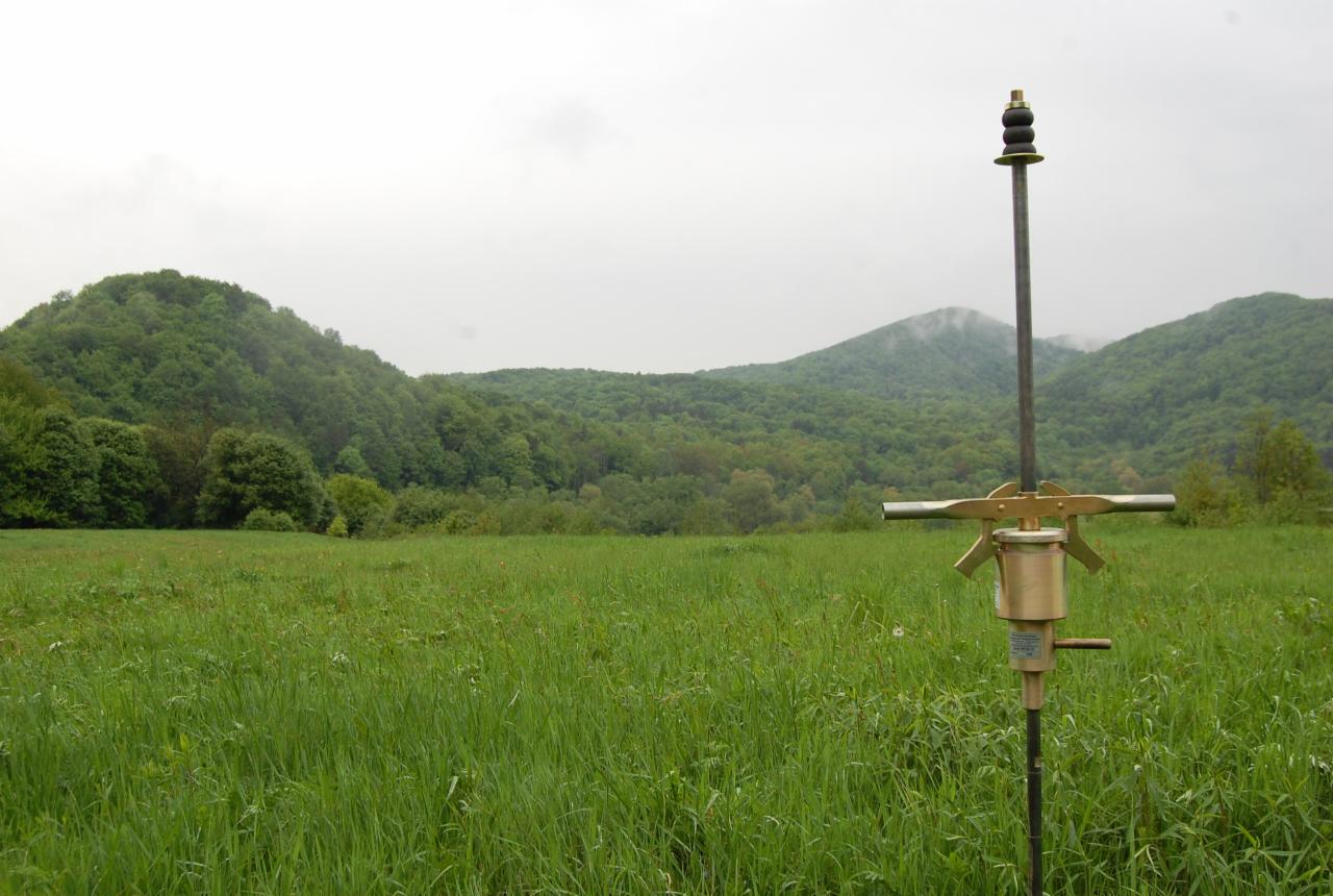 Urządzenie do pobierania próbek gruntu na łące, w tle zalesione wzgórza i pochmurne niebo.