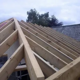 Konstrukcja drewniana dachu z widocznymi krokwiami, na tle częściowo pokrytego dachu z kominem i pochmurnego nieba.