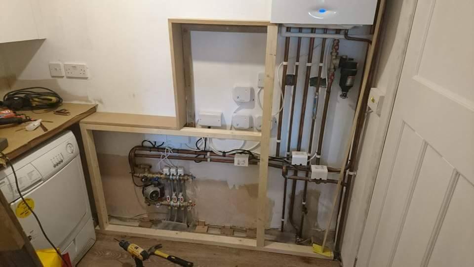 Montaż instalacji grzewczej w pomieszczeniu gospodarczym, widoczne rury miedziane, rozdzielacz, piec gazowy, obudowa z surowego drewna, pralka i narzędzia.