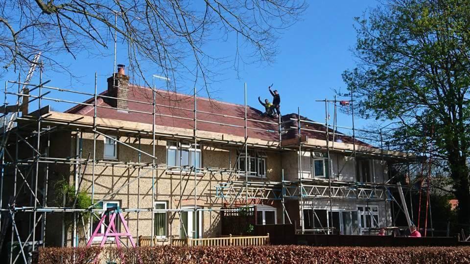 Dom jednorodzinny w trakcie wymiany dachu, otoczony rusztowaniem, dwaj robotnicy na dachu wznoszą ręce w geście radości.
