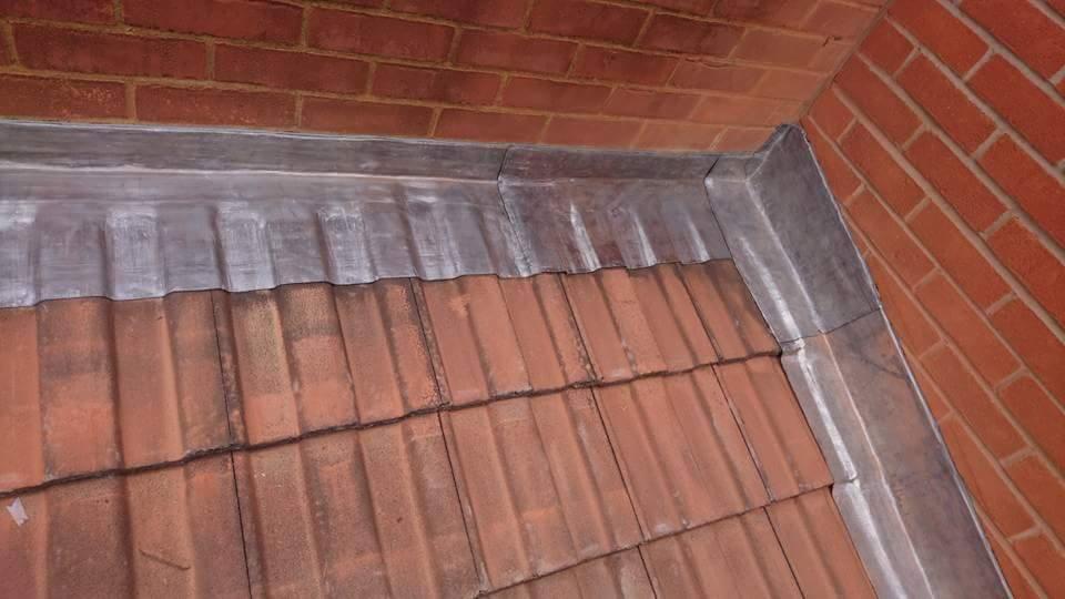 Szczegółowe ujęcie połączenia dachu z ceglaną ścianą, obróbka blacharska z ołowiu, widoczne ślady patyny na metalu i zabrudzenia na czerwonej dachówce.
