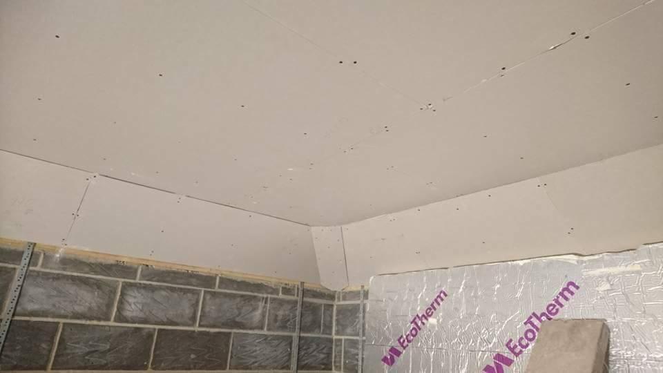 Sufit i ściana w trakcie prac wykończeniowych, widoczne płyty kartonowo-gipsowe mocowane do konstrukcji, częściowo ocieplone folią z nadrukiem EcoTherm, ślady szpachlowania na łączeniach płyt.