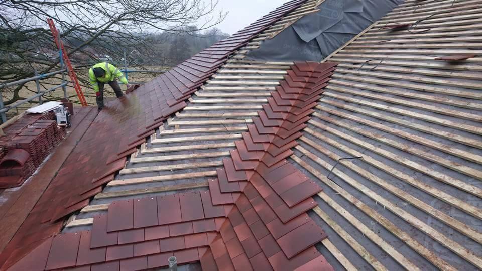 Montaż ciemnoczerwonej dachówki karpiówki na drewnianej konstrukcji dachu, widoczny dekarz w żółtej kurtce i stosy dachówek, tło stanowią drzewa bez liści.