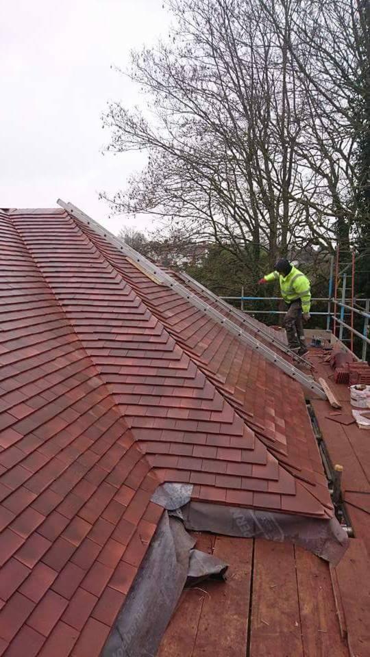 Czerwony dach z dachówki ceramicznej w trakcie układania, robotnik w jaskrawej kurtce na drabinie, widoczne drzewa w tle.