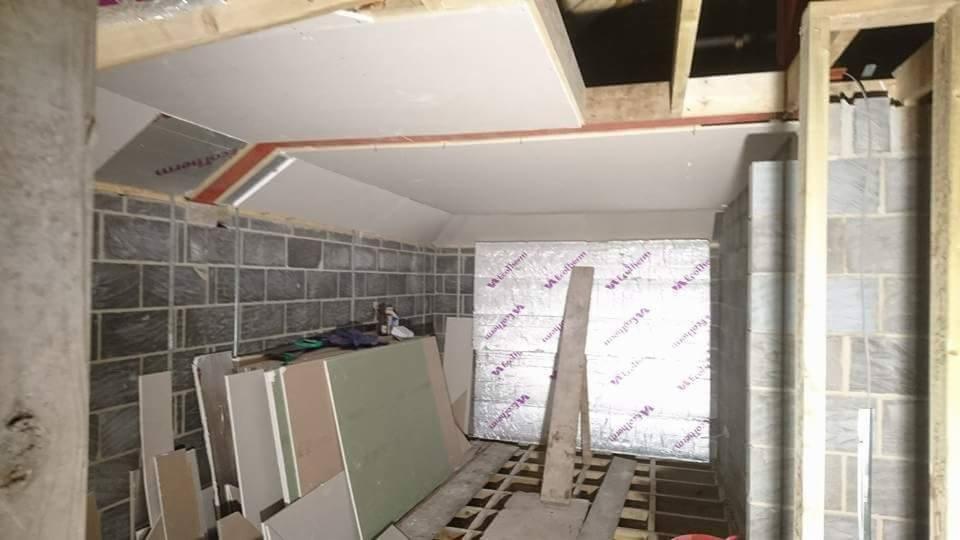 Wnętrze w trakcie prac wykończeniowych, widoczne surowe ściany z szarej cegły, częściowo zamontowana izolacja z folii aluminiowej, płyty gipsowo-kartonowe czekające na montaż i drewniana...