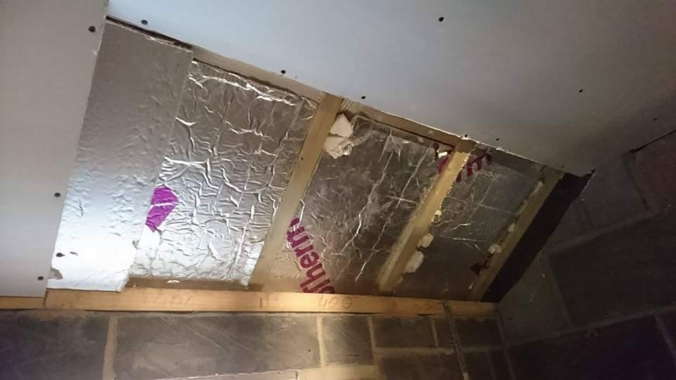 Skośny sufit w trakcie wykończenia: widoczne drewniane belki, izolacja z folią aluminiową z fragmentami różowej etykiety, częściowo zamontowana płyta gipsowo-kartonowa z widocznymi śrubami.