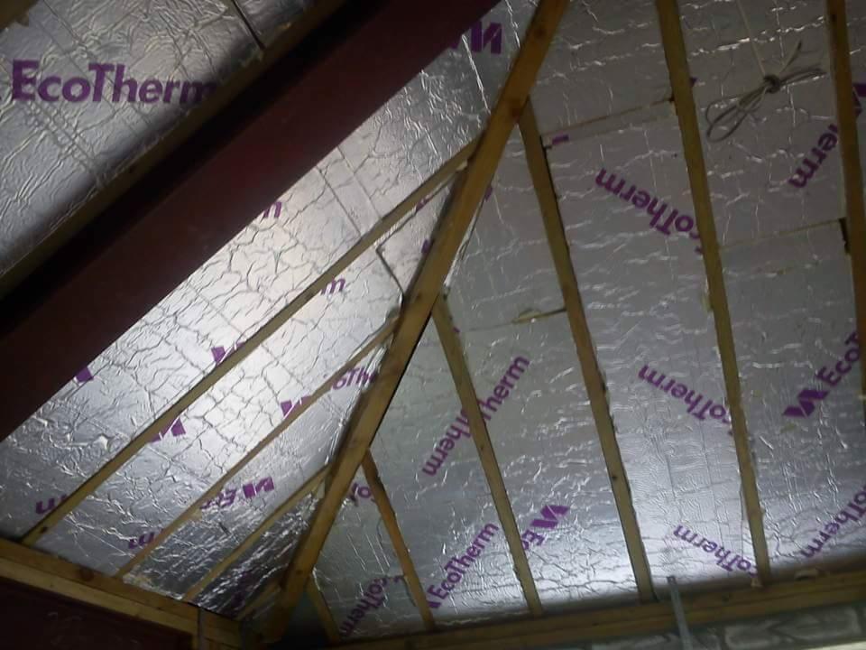 Izolacja poddasza z folią aluminiową i drewnianą konstrukcją więźby dachowej, widoczne logo EcoTherm.