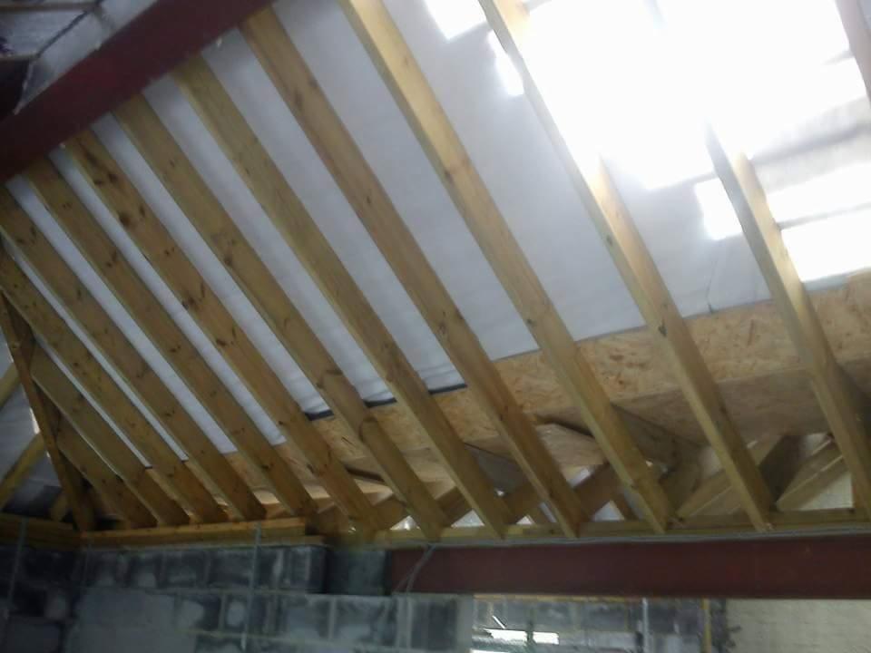 Konstrukcja drewniana dachu z membraną w trakcie montażu, widok od wewnątrz budynku, z widocznymi elementami muru i stalowymi belkami nośnymi.