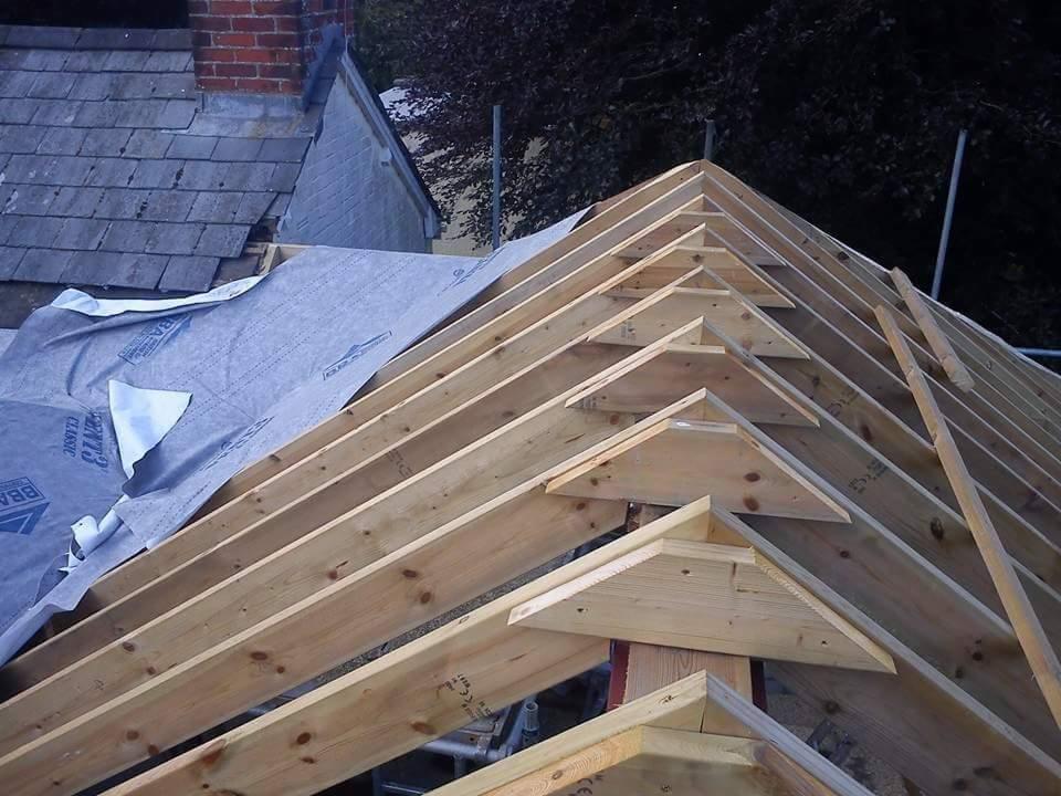 Konstrukcja drewniana dachu w trakcie budowy, widoczne krokwie i membrana dachowa, w tle fragment dachu z szarej dachówki i komin z czerwonej cegły.