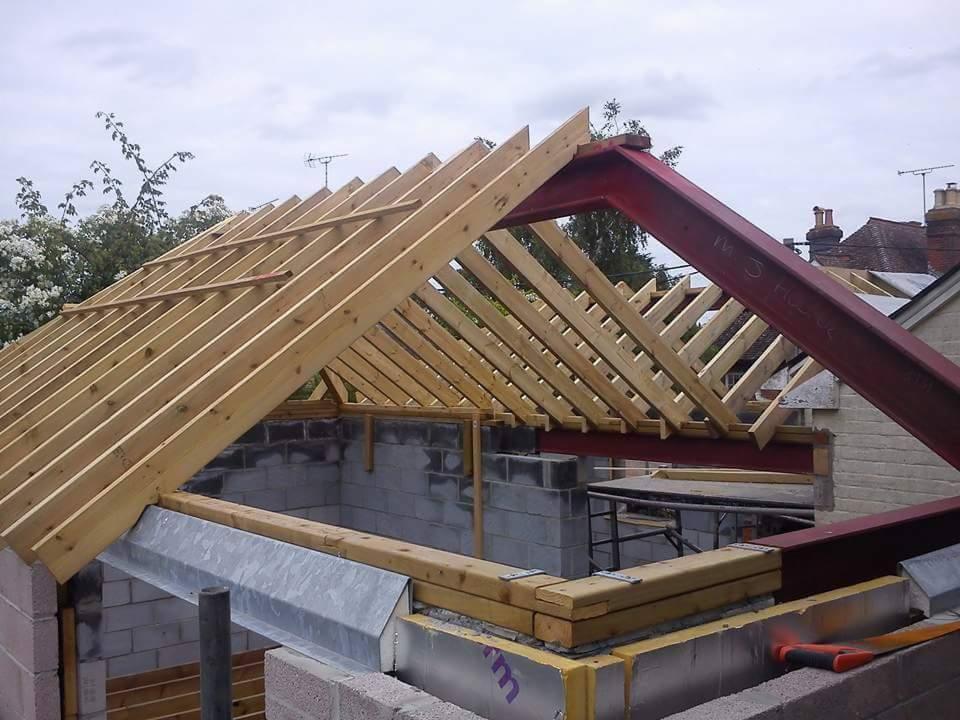 Konstrukcja więźby dachowej z drewnianych krokwi i czerwonej belki stalowej, widoczne bloczki betonowe i izolacja, etap budowy domu.