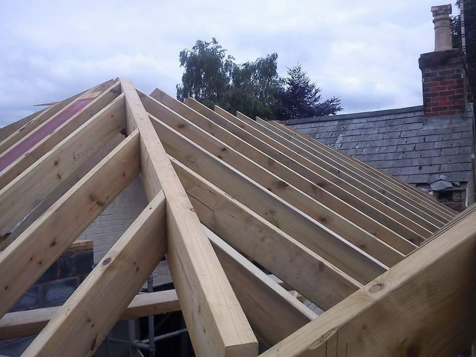 Konstrukcja drewniana dachu z widocznymi krokwiami, na tle częściowo pokrytego dachu z kominem i pochmurnego nieba.