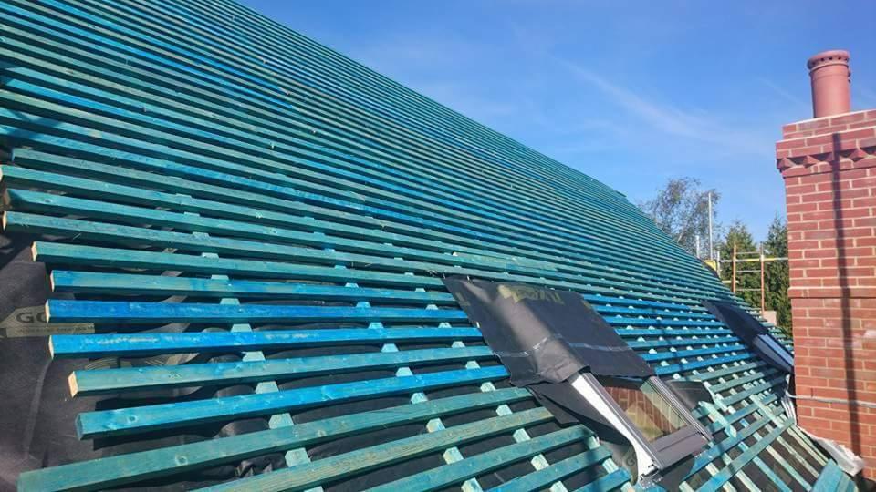 Niebieskie łaty dachowe pod dachówkami, widoczne okna dachowe z czarnym zabezpieczeniem oraz ceglany komin na tle błękitnego nieba.