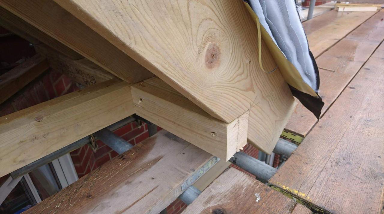 Fragment drewnianej konstrukcji dachu podczas montażu z widocznymi elementami więźby, deskowaniem i fragmentem rusztowania.