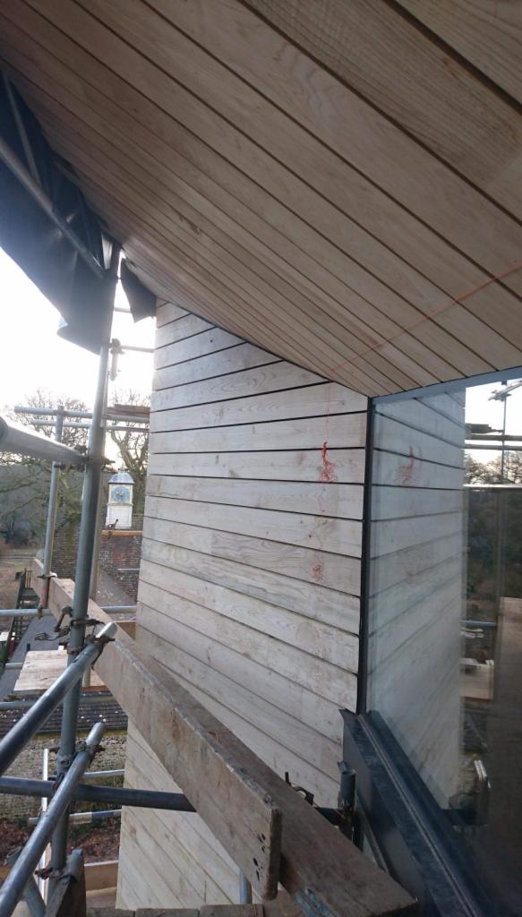 Montaż elewacji drewnianej z desek na budynku, widoczne rusztowanie i okno, jasne drewno, częściowo pod zadaszeniem.