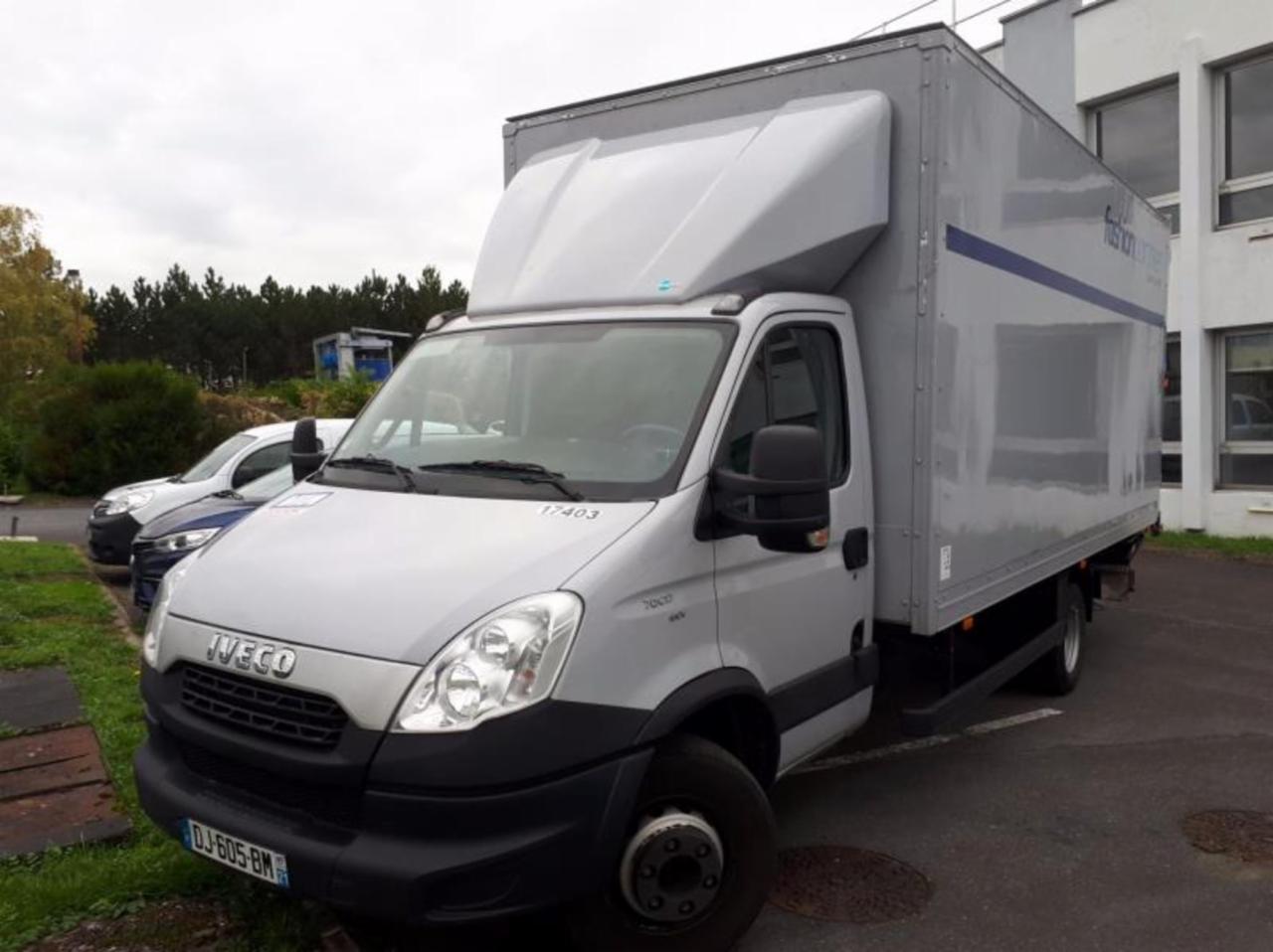 Szary samochód dostawczy Iveco z zabudową kontenerową, zaparkowany na parkingu przed budynkiem, z widocznymi innymi samochodami w tle.