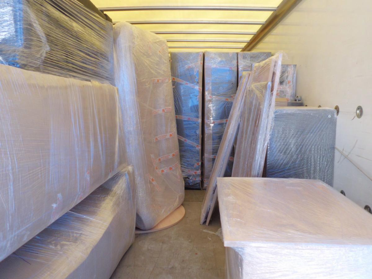 Wnętrze busa wypełnione meblami owiniętymi folią stretch, przygotowanymi do transportu. Widoczne zabezpieczone krzesła, szafy i inne elementy wyposażenia wnętrz.