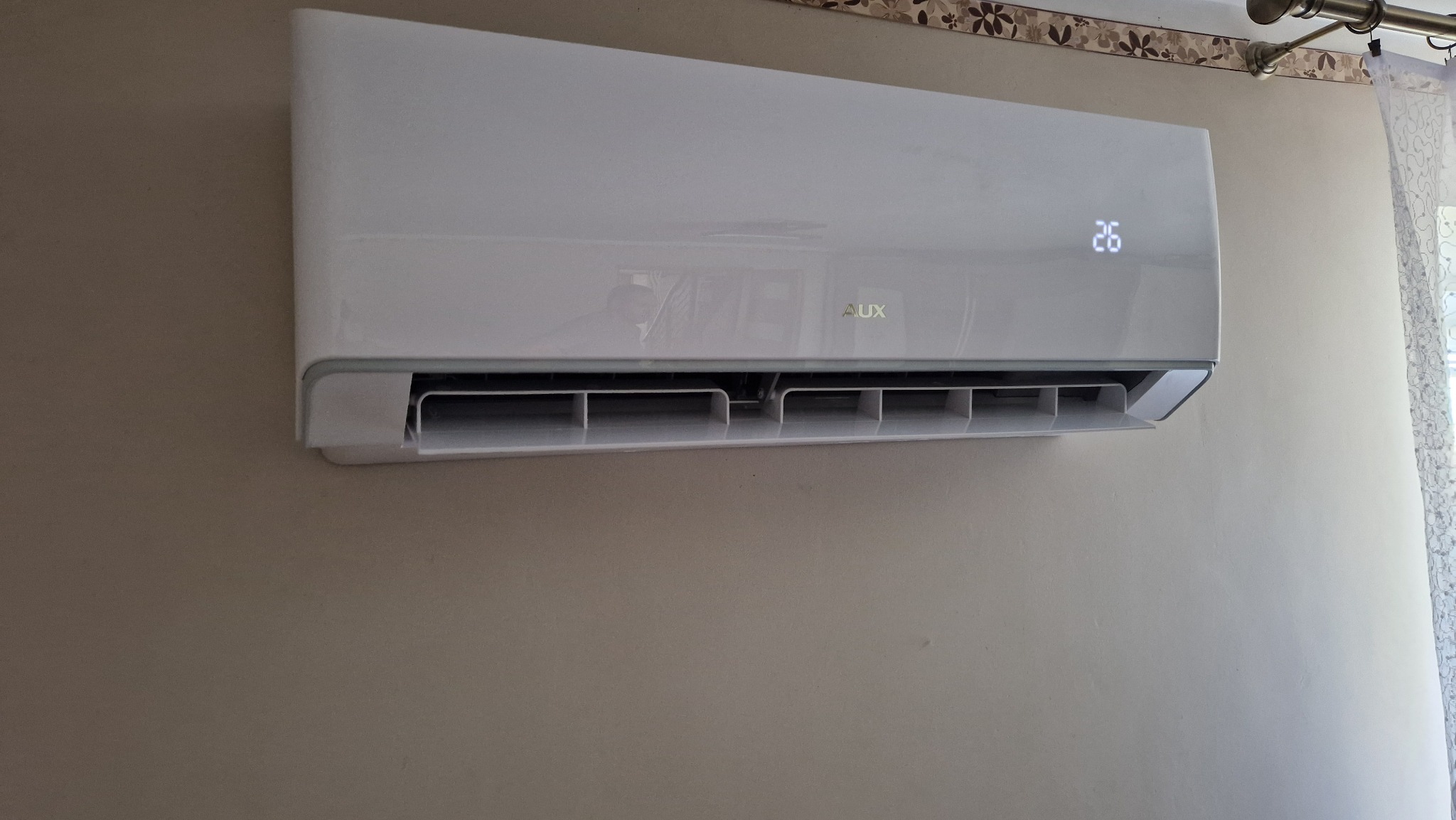 Biała jednostka klimatyzacji AUX zamontowana na beżowej ścianie w pokoju z widoczną zasłoną w tle, wyświetlająca temperaturę 26 stopni.