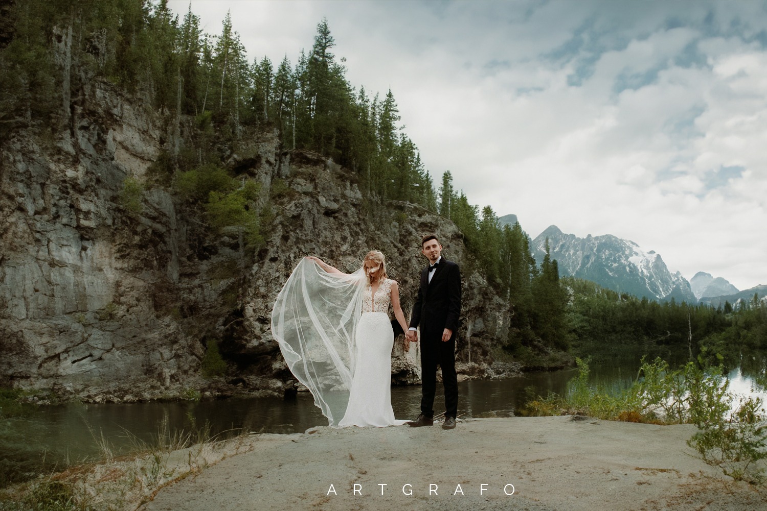 Para młoda, ona w białej sukni z koronkową górą i rozwianym welonem, on w czarnym garniturze z muszką, pozują na tle skalnej ściany, jeziora i gór.
