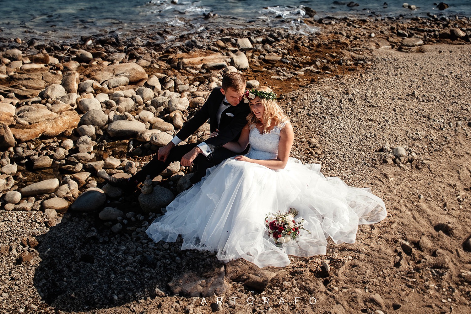 Para młoda siedzi na kamienistej plaży nad brzegiem morza; panna młoda w białej sukni z wiankiem na głowie trzyma bukiet, pan młody w czarnym garniturze opiera się o kamienie.