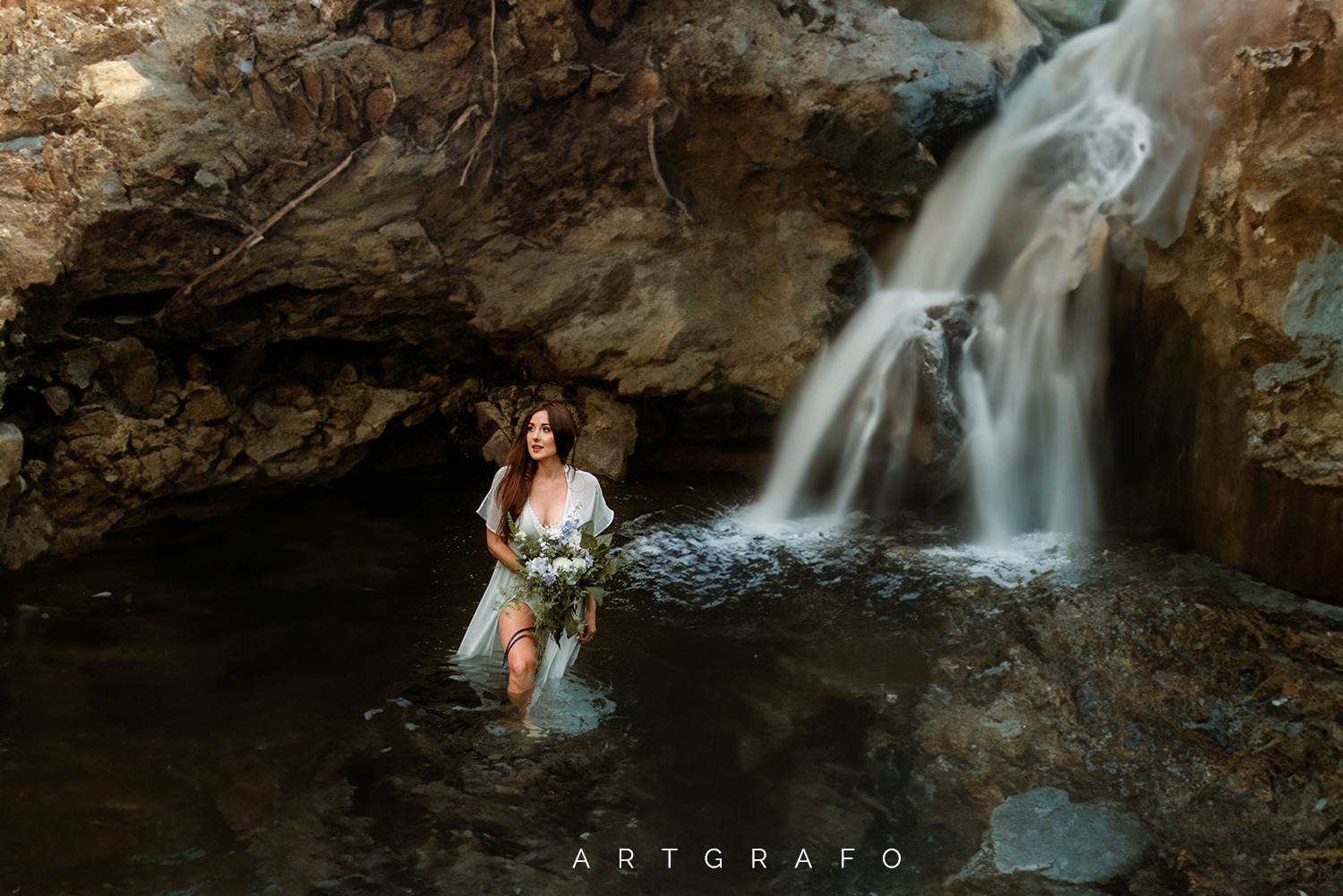 Kobieta w zwiewnej sukience i z bukietem stoi w wodzie pod wodospadem w skalnej grocie.