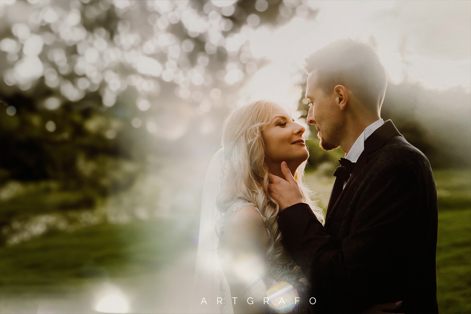 Para młoda w zbliżeniu, panna młoda z blond włosami i welonem, pan młody w czarnym smokingu z muszką, delikatne światło słoneczne, romantyczna atmosfera.