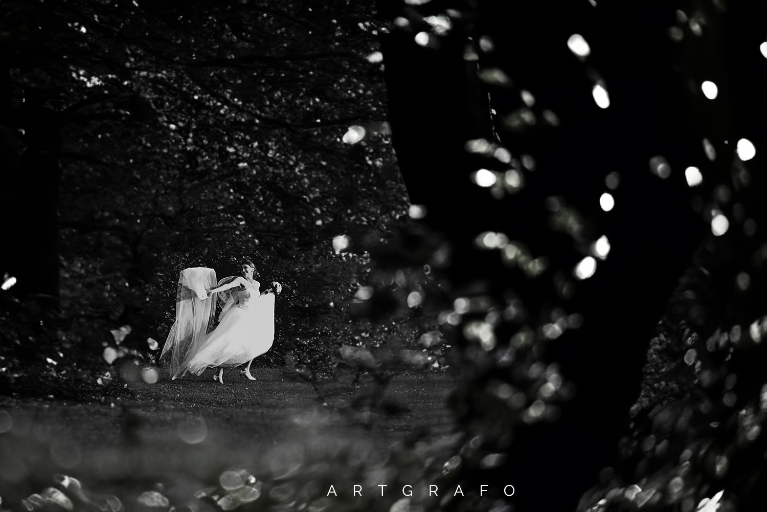 Czarno-białe ujęcie panny młodej w zwiewnej sukni, trzymającej bukiet, w ruchu na tle ciemnego lasu, z efektem bokeh w pierwszym planie.