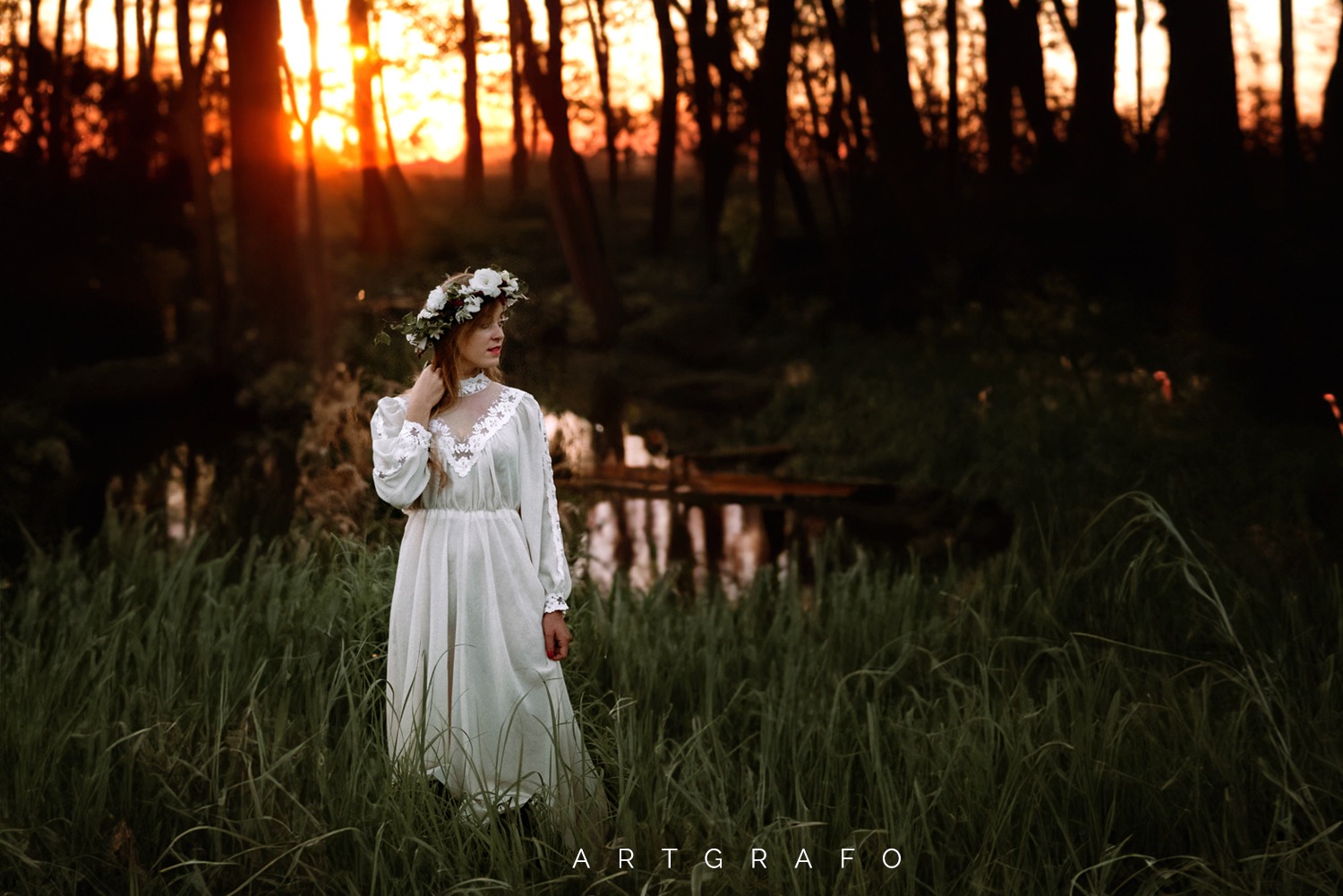 Kobieta w długiej, białej sukni z koronkowymi detalami i wiankiem z białych kwiatów na głowie, pozuje w wysokiej trawie na tle leśnego krajobrazu o zachodzie słońca.