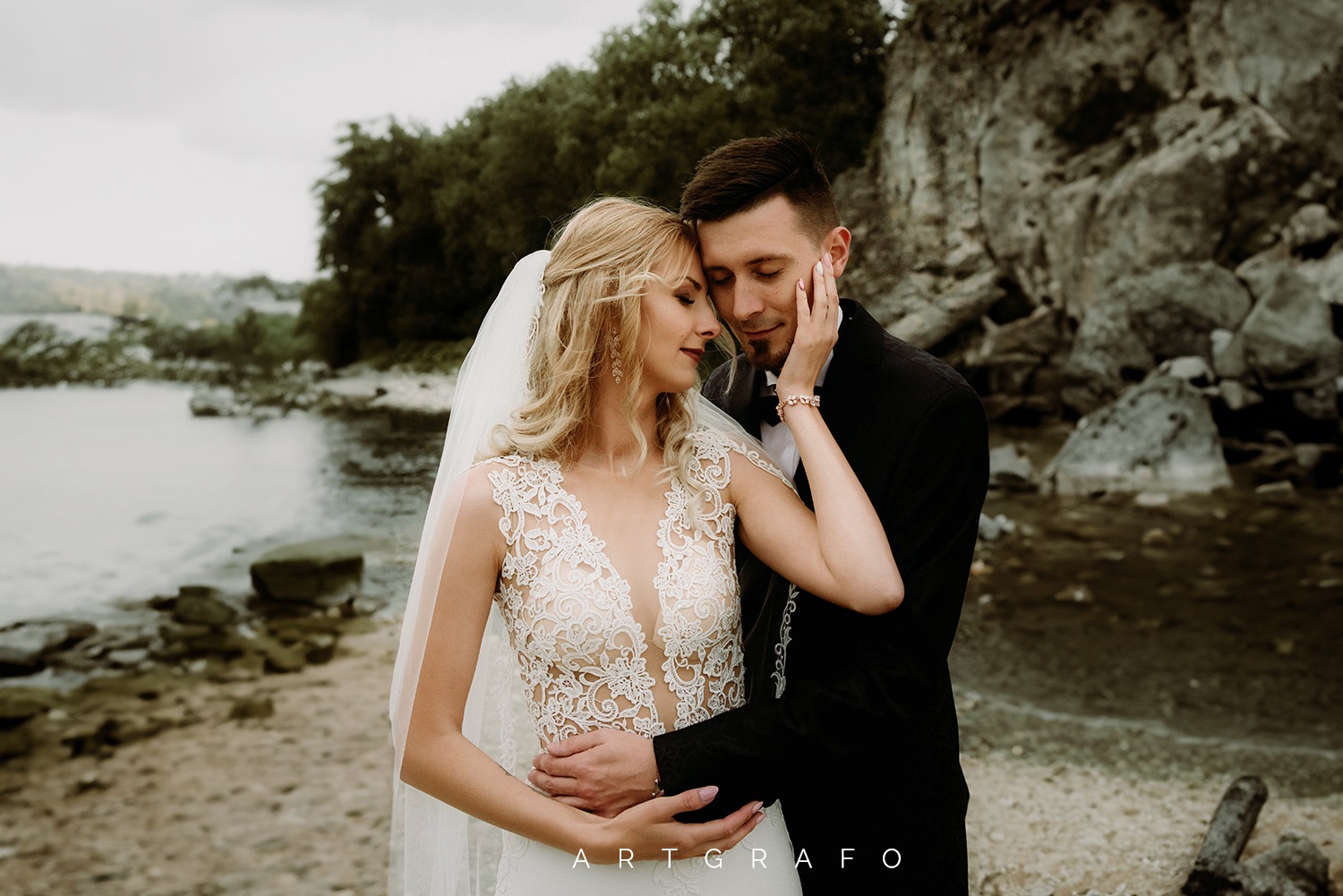 Para młoda w objęciach na tle jeziora i skał; panna młoda w koronkowej sukni z welonem, pan młody w czarnym smokingu z muszką.