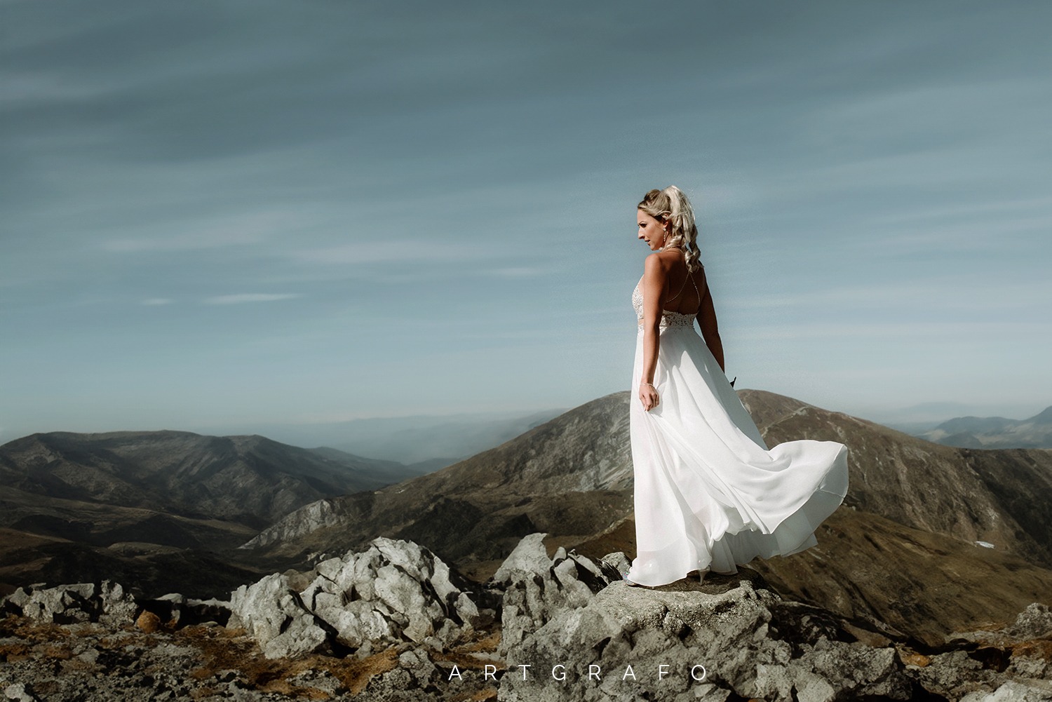 Panna młoda w zwiewnej białej sukni, stojąca na skalistym szczycie góry z rozległym widokiem na pasma górskie w tle.