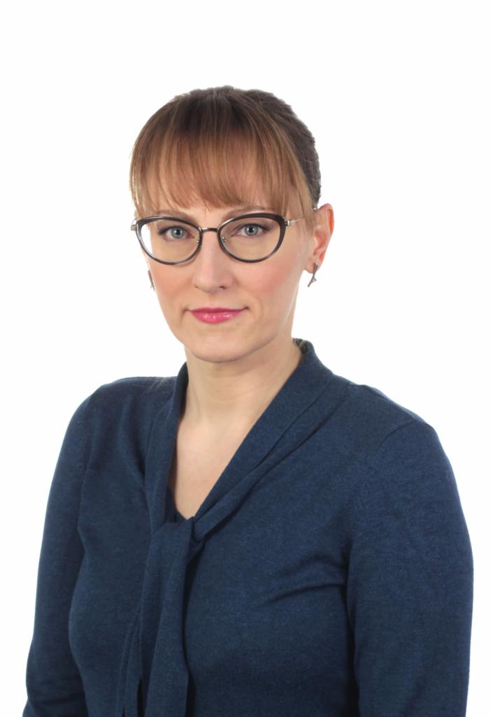 Portret kobiety w okularach, z grzywką, ubranej w ciemnoniebieską bluzkę z wiązaniem pod szyją, na białym tle.