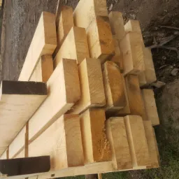 Stos świeżo przetartej, jasnej tarcicy ułożonej na palecie na tle ziemi i drzew, widoczny przekrój belek z wyraźnymi słojami.