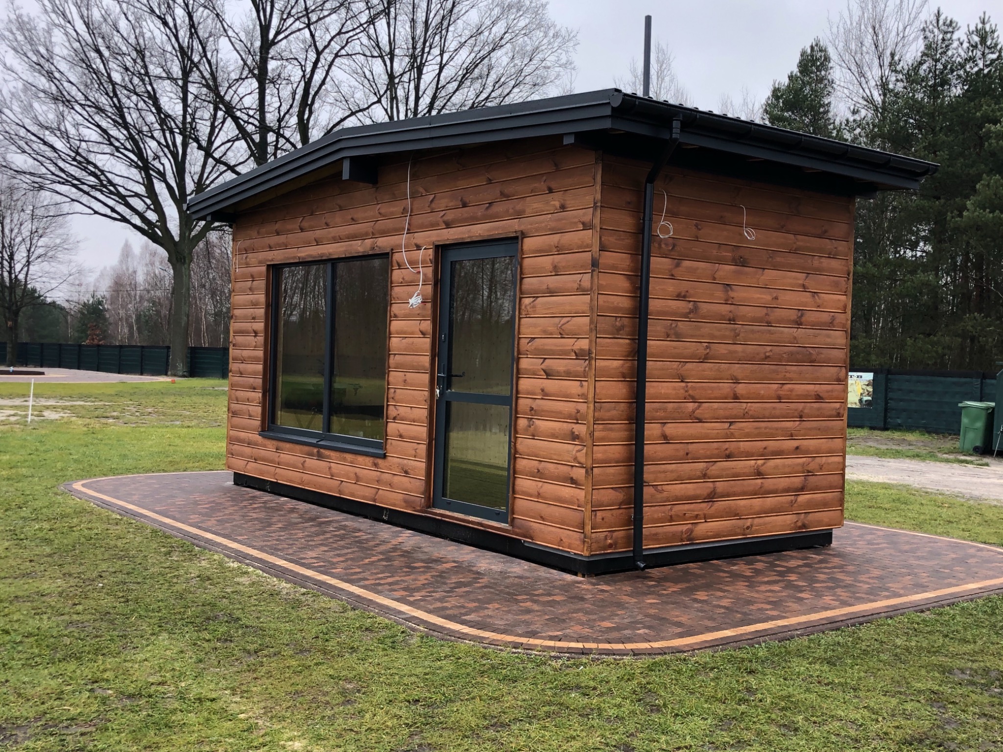 Gotowy drewniany domek z czarną stolarką i dachem, posadowiony na kostce brukowej w ogrodzie. Widoczne rury spustowe i przewody elektryczne.