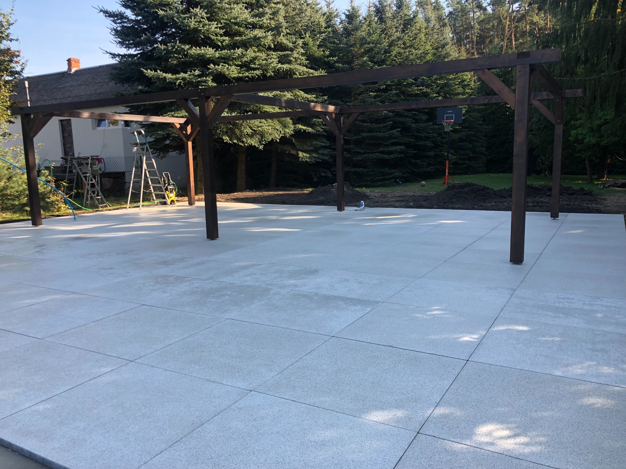 Nowoczesna, ciemnobrązowa konstrukcja drewniana zadaszenia tarasu na jasnej, kwadratowej, betonowej płycie, w tle dom i wysokie drzewa iglaste.