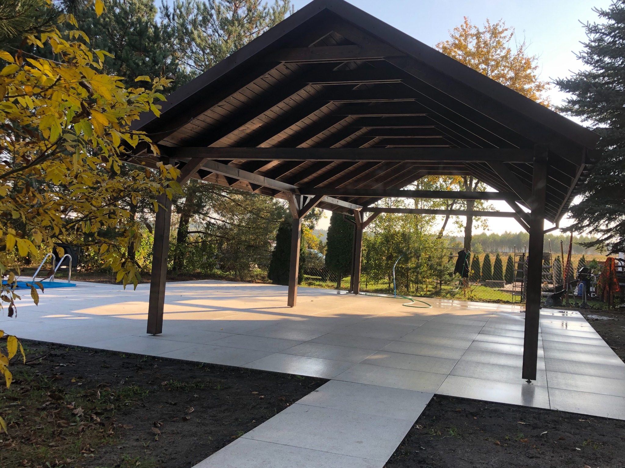 Drewniana altana z ciemnym dachem, płytki tarasowe w jasnym odcieniu, widok na ogród z basenem w tle, jesienne liście na drzewie po lewej stronie.