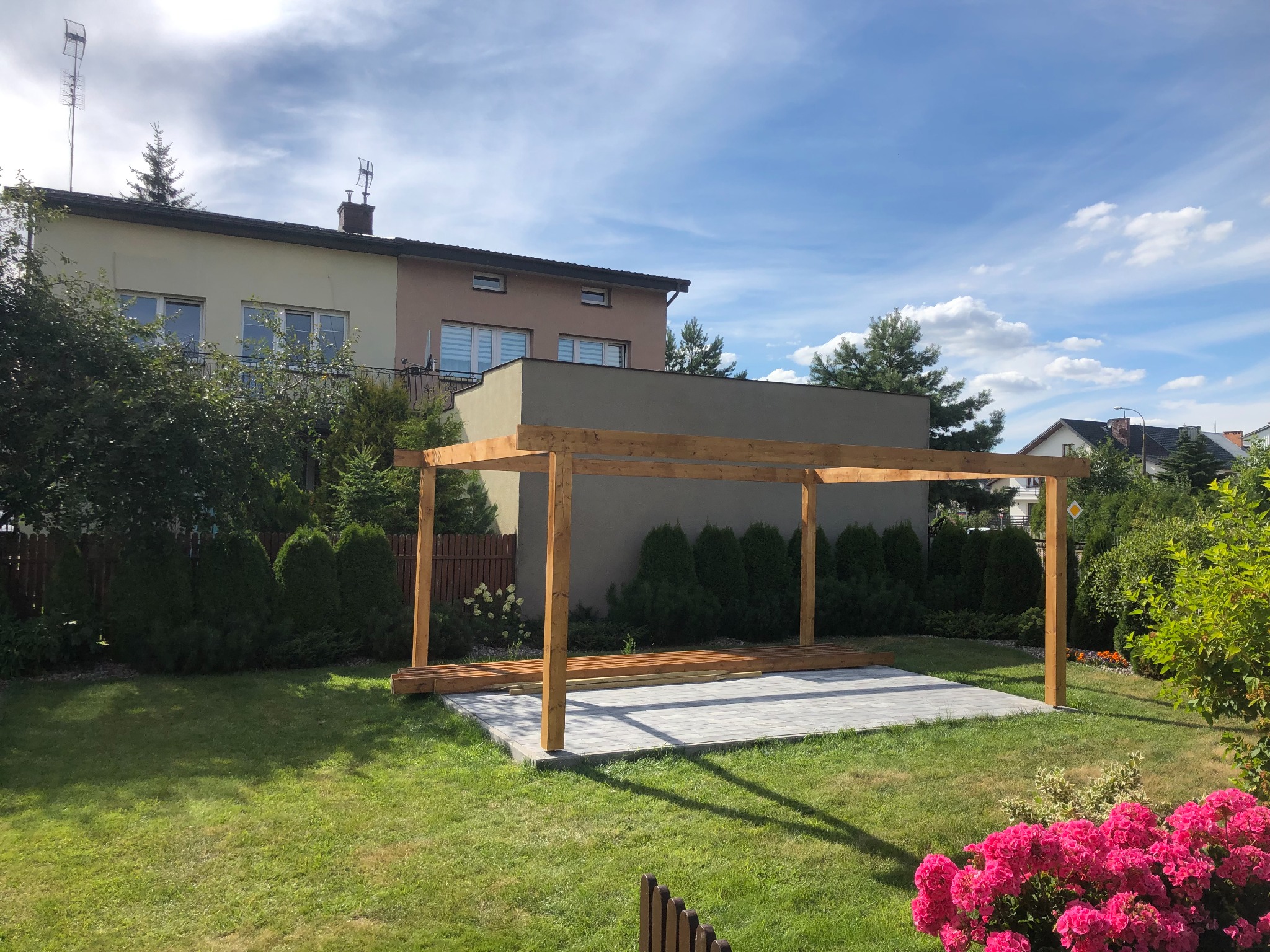 Drewniana konstrukcja zadaszenia tarasu na wyłożonej kostką powierzchni, widok z ogrodu w słoneczny dzień.