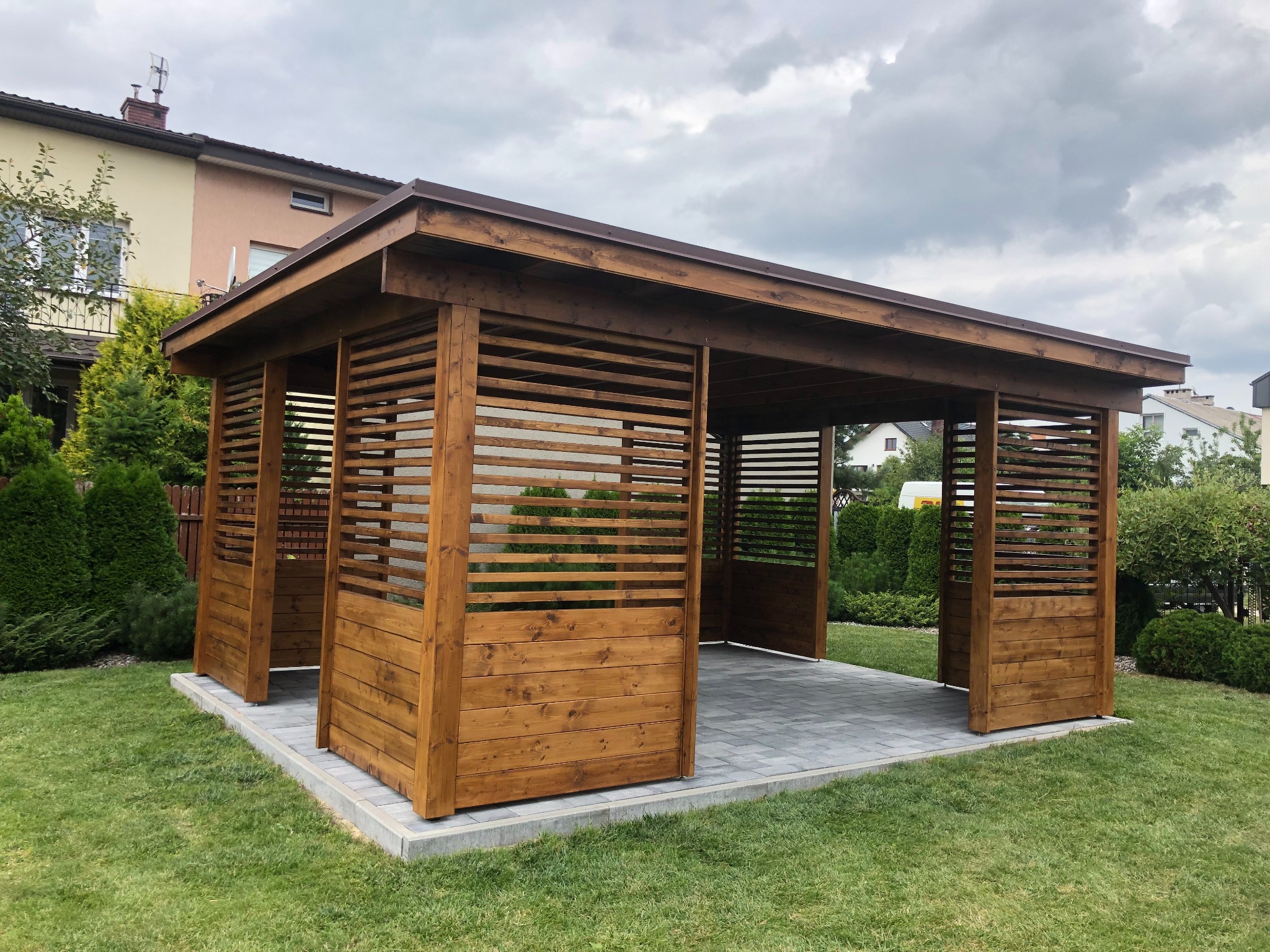 Drewniana altana z poziomymi deskami, zbudowana na szarej kostce brukowej, otoczona zielonym trawnikiem i tujami w ogrodzie.