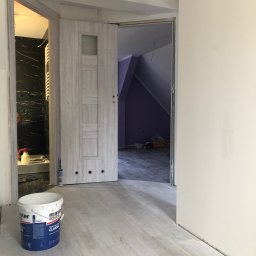 S-BAU USŁUGI OGÓLNOBUDOWLANE STANKIEWICZ - Wnętrze w trakcie remontu: widok na łazienkę z czarną marmurową ścianą i otwarte drzwi do poddasza z fioletową ścianą, na pierwszym planie wiadro z farbą Master Classic na jasnej podłodze.