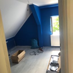 S-BAU USŁUGI OGÓLNOBUDOWLANE STANKIEWICZ - Poddasze po malowaniu: ściany w odcieniach bieli i intensywnego błękitu, okno z widokiem na zieleń, grzejnik, kartony i narzędzia na podłodze z paneli.