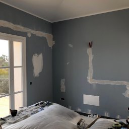 S-BAU USŁUGI OGÓLNOBUDOWLANE STANKIEWICZ - Przygotowania do malowania ścian w pokoju: zakryte folią meble, ściany z zagruntowanymi ubytkami, widoczne puszki elektryczne i narzędzia. Świeże światło wpada przez okno balkonowe.