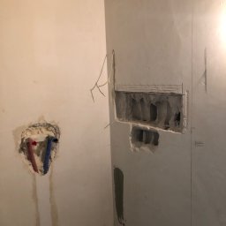 S-BAU USŁUGI OGÓLNOBUDOWLANE STANKIEWICZ - Ściana w trakcie przygotowania do instalacji, widoczne wycięte otwory na rury i instalacje, zaznaczone linie pionowe, pozostałości gruzu na podłodze.