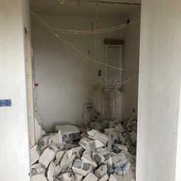 S-BAU USŁUGI OGÓLNOBUDOWLANE STANKIEWICZ - Wnętrze w trakcie remontu: sterta gruzu z pustaków na podłodze, odsłonięte przewody elektryczne na ścianie, widoczny grzejnik drabinkowy, ślady po instalacji na ścianie.
