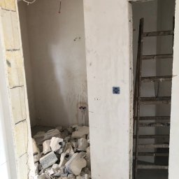 S-BAU USŁUGI OGÓLNOBUDOWLANE STANKIEWICZ - Wnętrze w trakcie prac: gruz po wyburzeniach, odsłonięte przewody elektryczne, stara drewniana drabina oparta o ścianę w przejściu.