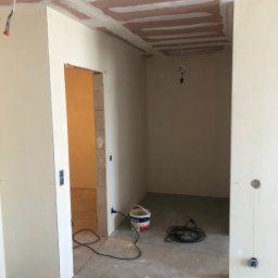 S-BAU USŁUGI OGÓLNOBUDOWLANE STANKIEWICZ - Wnętrze w trakcie prac wykończeniowych: sufit z płyt kartonowo-gipsowych z widocznymi łączeniami, ściany przygotowane do malowania, widoczne przewody elektryczne i wiadro z farbą Dekoral na podłodze.